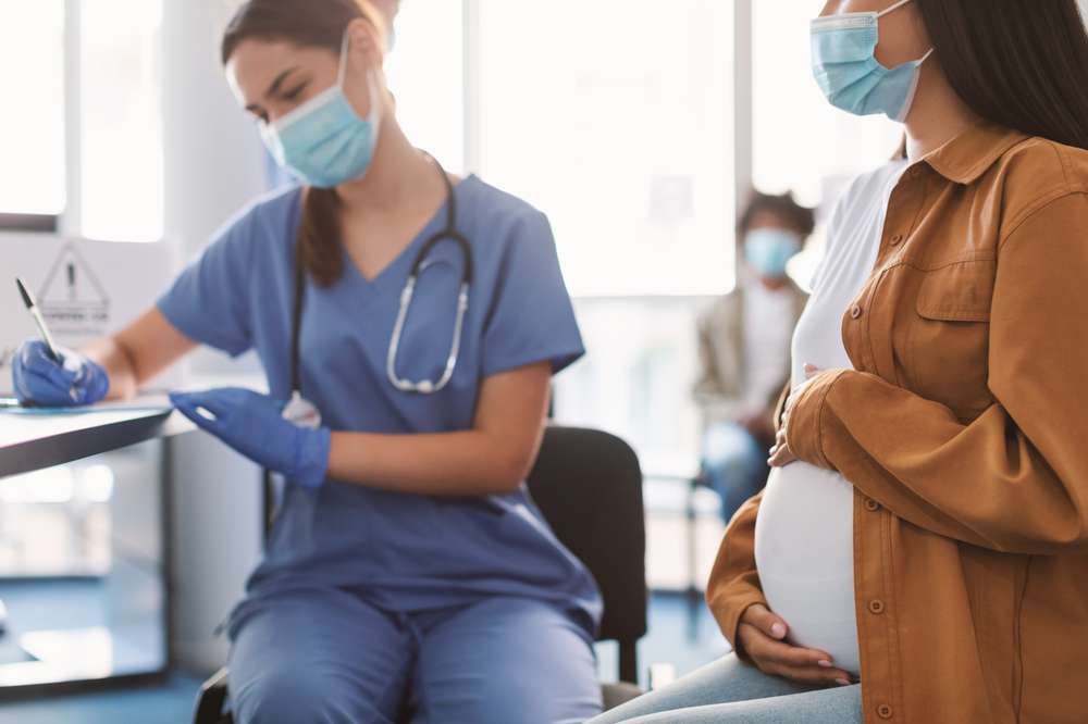 Mulher grávida com máscara facial visita ginecologista em hospital ou clínica médica para consulta, o médico preenche o registo de saúde, foco seletivo no abdómen. Controlo de saúde materna. Conceito de ginecologia.
