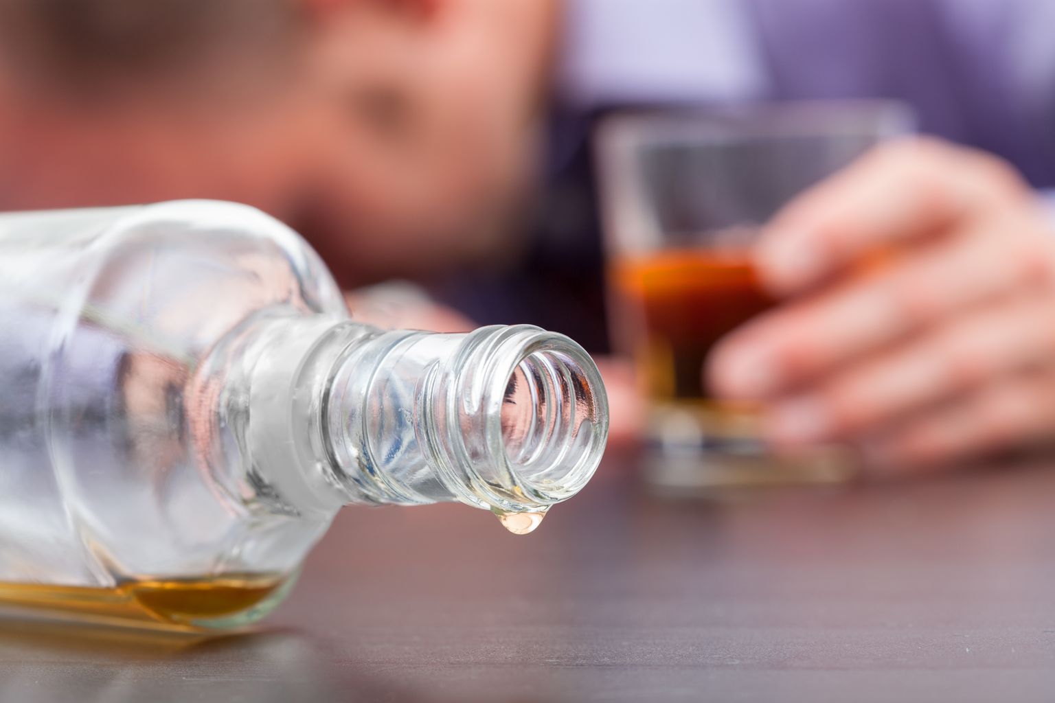 Primer plano de una botella de whisky caída y casi vacía con una persona dormida con un vaso de whisky medio lleno al fondo.