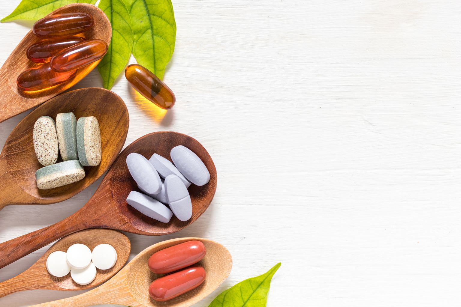 Varietà di pillole di vitamina in cucchiai di legno su sfondo bianco con foglia verde, prodotto complementare e medico, superficie piana profana