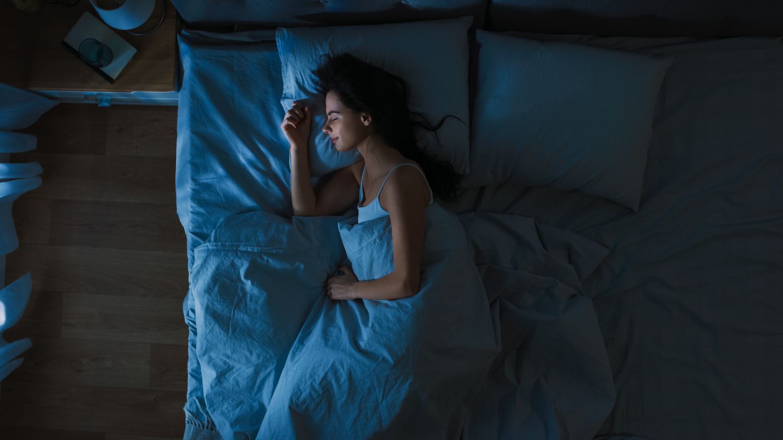 Top view of beautiful young woman sleeping comfortably on a bed in his bedroom at night. Blue night colors with cold weak lamppost light shining through the window.