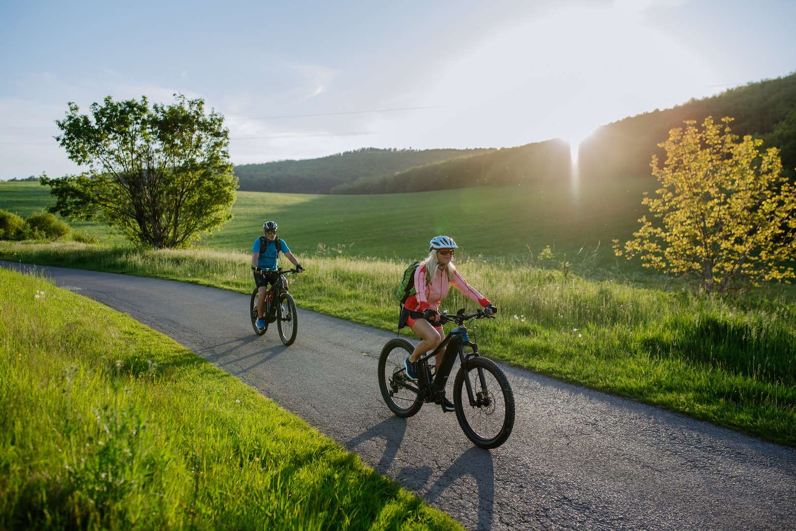 Active senior couple riding electric bicycles on road at summer park, healthy lifestyle concept. Aufnahme von zwei radfahrenden Personen in einer sommerlichen Landschaft.