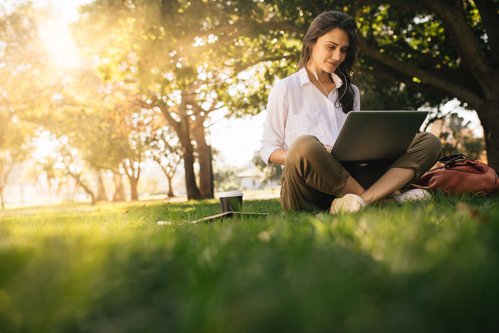 Een vrouw werkt buiten op een laptop met bomen op de achtergrond. Vrouw zittend op gras in park werkend aan laptop. Vrouwen die een hoofdtelefoon met laptop dragen terwijl ze onder een boom in een park zitten met fel zonlicht van achteren.