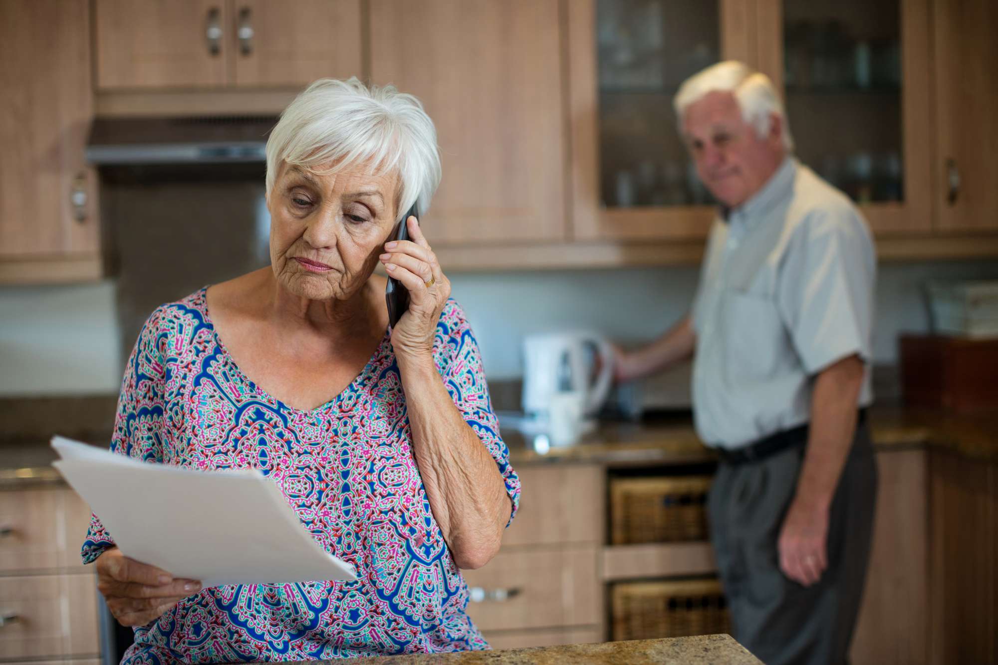 Mulher mais velha ao telefone com uma nota na mão, homem mais velho no fundo