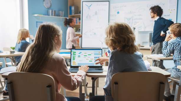 Une classe avec deux enfants au premier plan. Ils fonctionnent avec un ordinateur portable. Classe de sciences à l'école primaire : Un petit garçon et une petite fille en bandoulière utilisent un ordinateur portable avec un écran montrant un logiciel de programmation. Un professeur de physique explique une leçon à une classe diversifiée d'enfants intelligents.