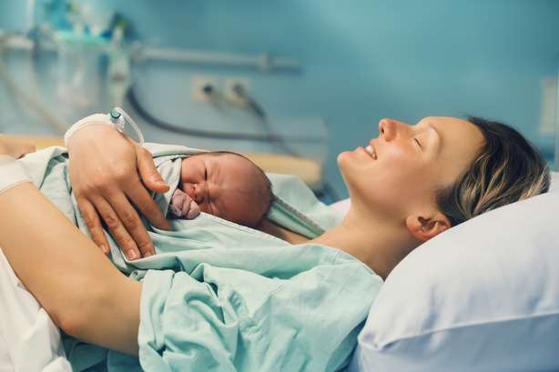 Mutter und Neugeborene. Geburt eines Kindes im Mutterschaftskrankenhaus. Junge Mutter umarmt ihr Neugeborenes nach der Geburt. Frauen gebären. Erste Momente des Babylebens nach der Arbeit. Eine Mutter haltet liegend ihr neugeborenes Kind auf ihrer Brust in einem Krankenhaus und lächelt dabei mit geschlossenen Augen.