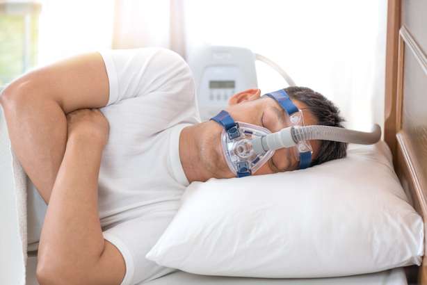 Fröhlicher und gesunder Senior Mann mit Cpap Maske schlafend die ganze Nacht lang auf seiner linken Seite Cross Arme ohne Schnarchen.Obstruktive Schlafapnoe Therapie. Ein älterer Herr trägt eine Atmungsmaske während er auf der Seite im Bett liegend versucht einzuschlafen.