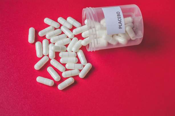 Weiße Tabletten in einem durchsichtigen, Placebo-gekennzeichneten Behälter. White placebo pills or capsules spilling out of a bottle on red background, placebo effect, randomization or treatment concept, vintage view