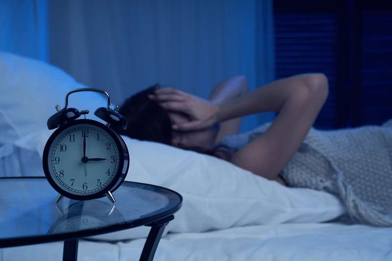 Uma mulher que sofre de insónias está na cama ao lado de um despertador. Mulher infeliz com insónias deitada na cama ao lado do despertador durante a noite