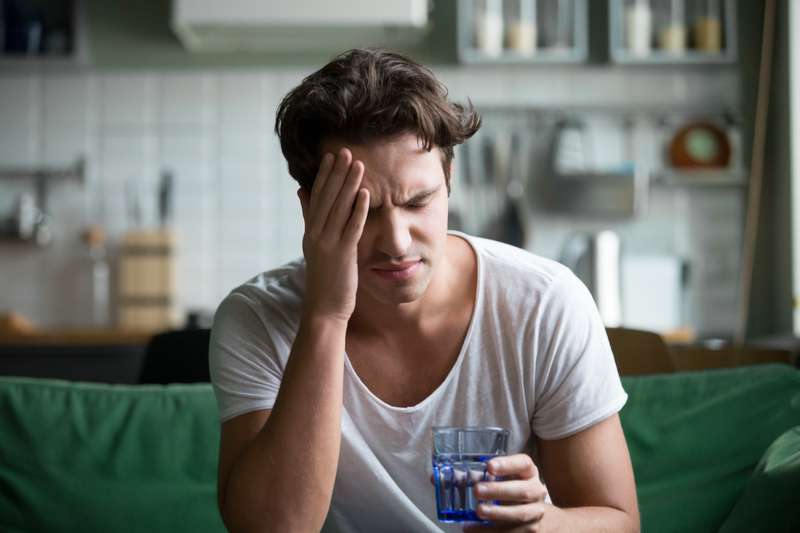 Jonge man heeft thuis last van hoofdpijn, migraine of een kater Shot van jonge man met hoofdpijn en een glas water in zijn hand